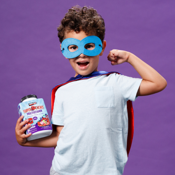 Young boy with a blue superhero mask flexing a bicep, as he holds Force Factor Kids Multivitamin Super Chews in the other hand.